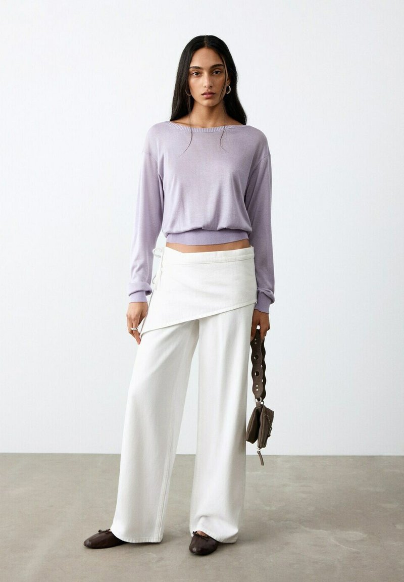 Mujer vistiendo una blusa de manga larga lavanda, pantalones blancos de pierna ancha con detalle de envoltura, zapatos planos marrones y sosteniendo un bolso marrón con correa en forma de lazo.