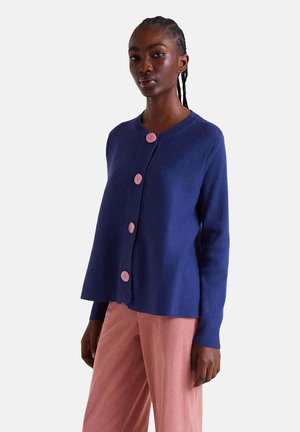 Cardigan azul marino fabricado con tejido suave, con cinco botones rosas en la parte delantera y un ajuste relajado. Combinado con pantalones de pana rosa.