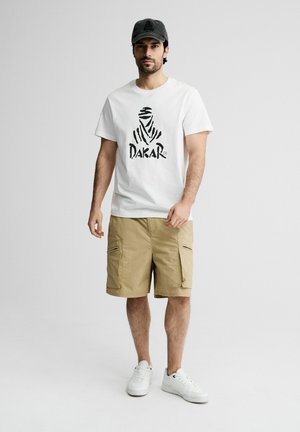Mężczyzna stojący, ubrany w biały t-shirt z logo Dakar, beżowe szorty cargo, białe trampki i ciemną czapkę na jednolitym szarym tle.