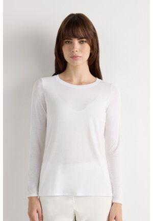 Frau mit braunen Haaren, die ein eng anliegendes weißes Langarmshirt und helle Hosen trägt, vor einem schlichten weißen Hintergrund.