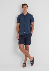 Short-sleeve navy button-up shirt, lightweight fabric, paired with navy shorts and dark sandals. Minimalist design with a relaxed fit.