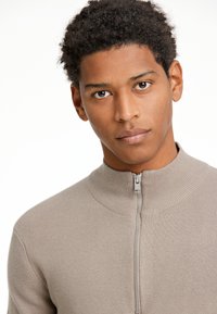 Beige neulepusero, jossa on korkea kaulus ja edessä vetoketju. Varustettu tekstuurilla ja istuvalla muotoilulla, joka korostaa modernia ilmettä.