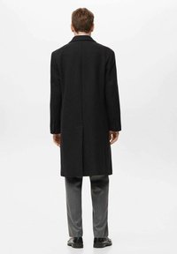 Langer schwarzer Wollmantel mit gerader Silhouette, Reverskragen und einer mittigen Rückenbelüftung, getragen über grauen Hosen und schwarzen Schuhen.