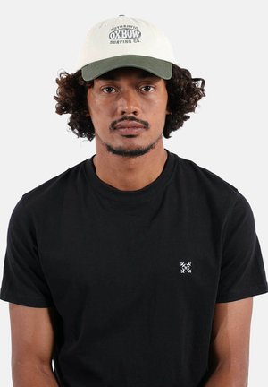 Homme aux cheveux bouclés portant un t-shirt noir et une casquette beige et verte avec le logo "Oxbow Surfing Co." sur un fond blanc.