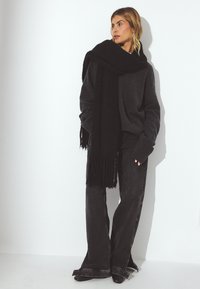 Oversize szary sweter z prążkowanymi mankietami, połączony z rozkloszowanymi czarnymi dżinsami. Długi czarny szalik opada na szyję, z frędzlami na końcach.