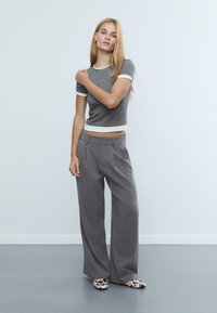 Grau Kurzarm-T-Shirt mit weißen Akzenten, kombiniert mit grauen weit geschnittenen Hosen. Das Modell trägt gemusterte Slipper. Neutrale Hintergrund.