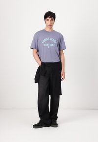 Violetinės medvilninės marškinėliai su užrašu "TOMMY JEANS NEW YORK" šviesiai mėlyna spalva, derinami su laisvais juodais džinsais ir juodais sportbačiais.