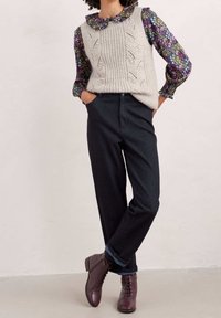 Strikket sweatervest i lys grå med kabelmønstre, lagdelt over en mørk blomsterbluse. Sammen med mørke jeans og brune snørebåndsstøvler.