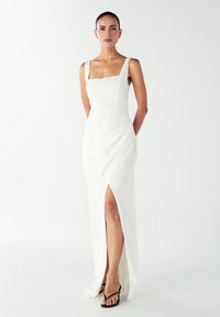 Žena u bele haljine bez rukava sa četvrtastim dekolteom i prorezom do butina, nosi crne sandale i stoji ispred obične bele pozadine.