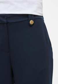 Close-up van marineblauwe broeken met een gestructureerde gouden knop op de riemlus, gedragen met een witte top.