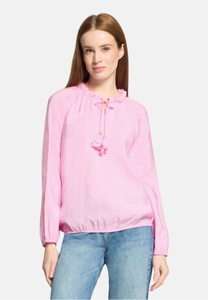 Young woman with straight red hair wearing a light pink long-sleeve blouse with a tied ruffled neckline and light blue jeans.