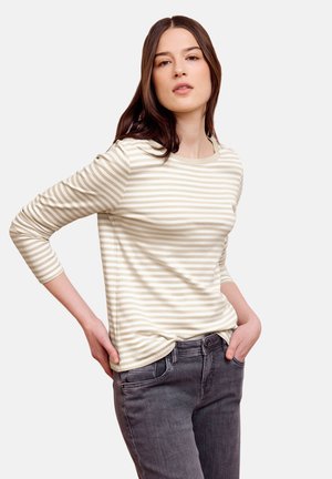 Junge Frau mit langen braunen Haaren, die ein beige-weiß gestreiftes Langarmshirt und dunkelgraue Jeans trägt, posiert mit den Händen in der Hüfte.