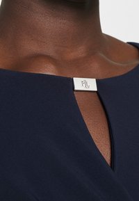 Närbild av en mörkhyad person som bär en marinblå topp med en liten silverklämma graverad med "RL" vid halsringningen.