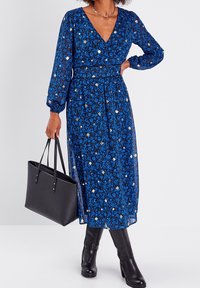 Robe à motif floral bleu avec de longues manches ballon, décolleté en V, taille cintrée et jupe fluide. Sac fourre-tout noir et bottes montantes.
