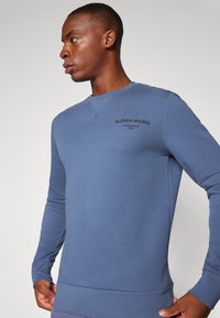 Sweatshirt azul com mangas longas, gola redonda e punhos canelados. Apresenta um logótipo subtil impresso no peito. Textura suave semelhante ao algodão.