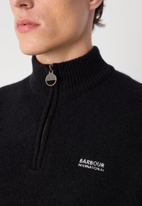 Sweater de fleece preto com colarinho alto canelado e zíper de um quarto. Apresenta um puxador de zíper de metal redondo e o logotipo branco "Barbour International".