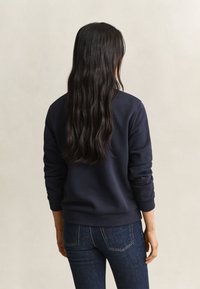 Vrouw met lang donker haar die een donkerblauwe trui en blauwe spijkerbroek draagt, met haar rug naar de camera tegen een effen lichte achtergrond.