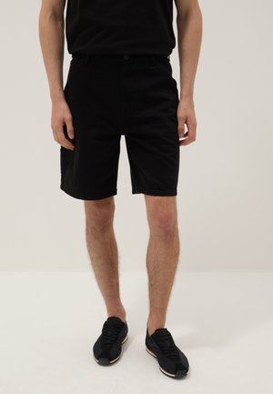 Hombre con pantalones cortos negros hasta la rodilla combinados con zapatillas negras, de pie frente a un fondo claro y liso.