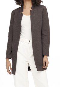 Abrigo marrón de manga larga con un diseño sin cuello, corte recto y tejido texturizado, combinado con una blusa blanca y pantalones de color claro.