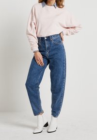 Ljust rosa sweatshirt med ribbade mudd över ett par blå denimbyxor med hög midja. Modellen bär vita ankelstövlar med klack.