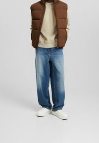 Brun pufferväst över en beige sweatshirt, kombinerad med lösa blå jeans och vita sneakers. Västet har hög krage och dragkedja.