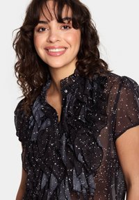 Črna prosojna bluza z nagubanimi detajli, z visokim ovratnikom in kratkimi rokavi, okrašena z zvezdnatim vzorcem.