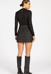 Vrouw met een bun en een zwart langemouwen top, een grijs geruite minirok en zwarte kniehoge laarzen, van achteren gezien.