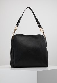 Černá kožená kabelka přes rameno s nastavitelným popruhem, kováním v zlaté barvě a hladkým texturovaným povrchem, vystavená na bílém podstavci.
