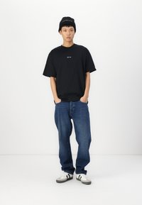 Svart t-skjorte med kort arm og hvit tekst, sammen med løse blå denimjeans og grå sneakers med svarte striper. Svart lue på hodet.