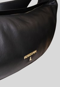 Sac à main en cuir noir avec une texture lisse, présentant un logo doré et un design simple et arrondi. Fermeture éclair en haut.