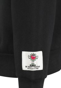 Schwarzer Pullover aus strukturiertem Stoff, mit gerippten Bündchen und einem weißen Etikett, das eine Cartoonfigur mit dem Text "BE HAPPY TOGETHER" zeigt.