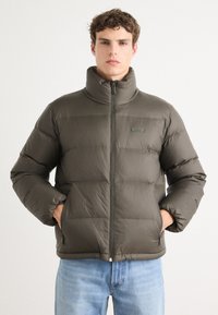 Jeune homme portant une veste matelassée zippée gris foncé avec un col montant et les mains dans les poches, assortie à un jean bleu clair.