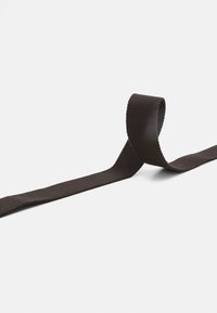 Brun vävd polyesterband, platt och flexibelt, med en slät yta. Krullat i ena änden, sträcker sig horisontellt mot en ljus bakgrund.