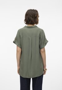 Blusa verde oliva con maniche corte arrotolate, tessuto leggero, dettaglio di pieghe sul retro e orlo leggermente arrotondato. Nessun motivo o accessorio visibile.