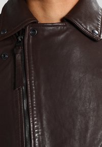 Bruine leren jas met een hoge kraag, ritssluiting en drukknopen. De textuur is soepel met een subtiele glans en gedetailleerde stiksels.