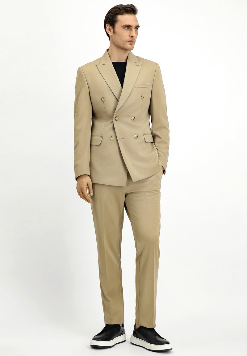 Completo beige doppiopetto con bottoni dorati, due tasche laterali e pantaloni abbinati. Indossato con sneakers nere, caratterizzate da una texture liscia.