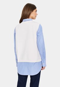 Weißer ärmelloser Pullover über einem hellblauen und weißen gestreiften Langarmshirt, kombiniert mit dunkelblauen Jeans. Sanfte Textur.