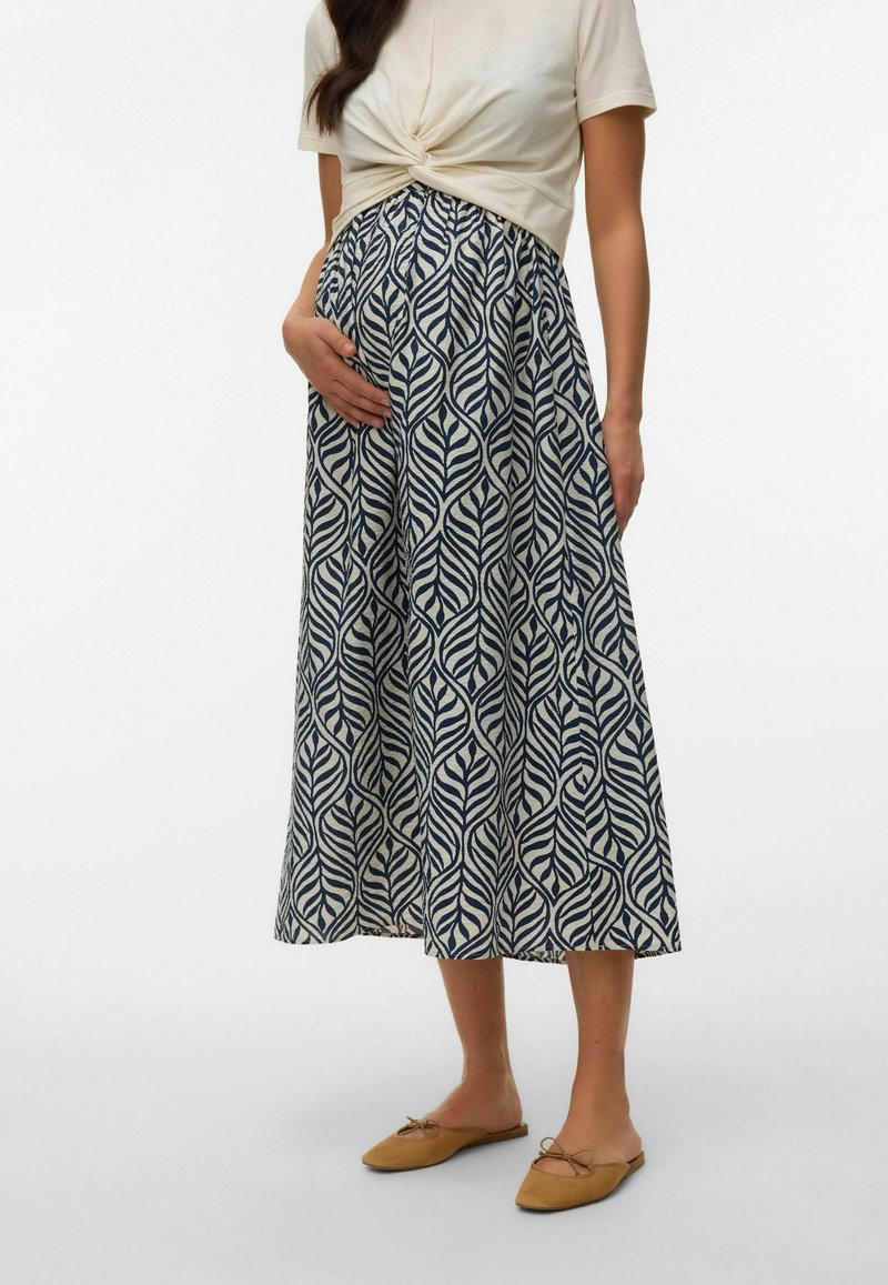 Femme enceinte portant un haut crème à manches courtes torsadé, une jupe à motifs bleus et blancs, et des ballerines camel, tenant son ventre.