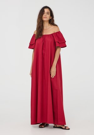 Abito lungo rosso scollato sulle spalle con maniche a strati e fluttuanti, silhouette fluida e tessuto morbido, abbinato a sandali neri.