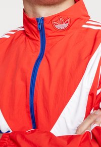 Röd vindjacka med vita detaljer och blå dragkedja. Har en upphöjd logotyp vid kragen och tre vita ränder på axlarna.