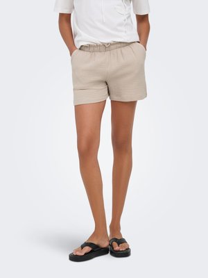 Woman wearing beige elastic waist shorts, white short-sleeve blouse, and black platform flip-flops standing with hands in pockets.