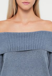 Απαλή μπλε πουλόβερ εκτός ώμου με ραβδωτό λαιμό και ομαλή υφή, χαρακτηριστικό χαλαρού στυλ και μινιμαλιστικής σχεδίασης.