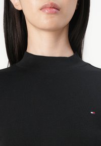 Crna rebrasta majica s visokim izrezom koja ima mali logotip u boji na prsima. Tkanina izgleda glatko s prilagodljivim siluetom.