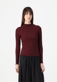 Långärmad ribbad topp i mörk burgundy med hög halsringning och rå kant, i kombination med en svart kjol för en lager-på-lager look.