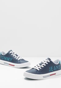 Navy blue sneakers made of synthetic material. Features white laces, with “TOMMY” and “JEANS” printed in teal. White rubber sole with red accents.