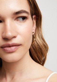 Boucles d'oreilles en or en forme de cerceau avec un design sculptural, mettant en avant un accent en pierre délicat. Finition métallique lisse, forme courbée et aspect léger.