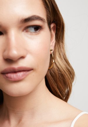 Pendientes de aro dorados con un diseño escultural, que presentan un delicado acento de piedra. Acabado metálico suave, forma curva y apariencia ligera.