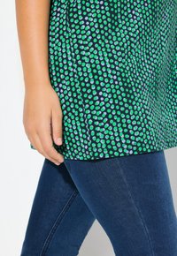 Top a pois verdi e blu con sfondo nero, caratterizzato da una vestibilità morbida e maniche corte, abbinato a jeans in denim scuro.