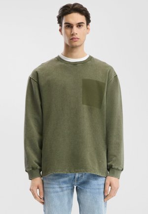 Zelena oversize sweatshirta od pamuka s okruglim izrezom, sadrži kvadratnu džepu na lijevoj strani grudnog koša, vidljive šavove i rebraste manžete.