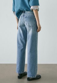 Person wearing light blue straight-leg jeans, black loafers, and a blue sweater over a striped shirt, standing on a gray floor.